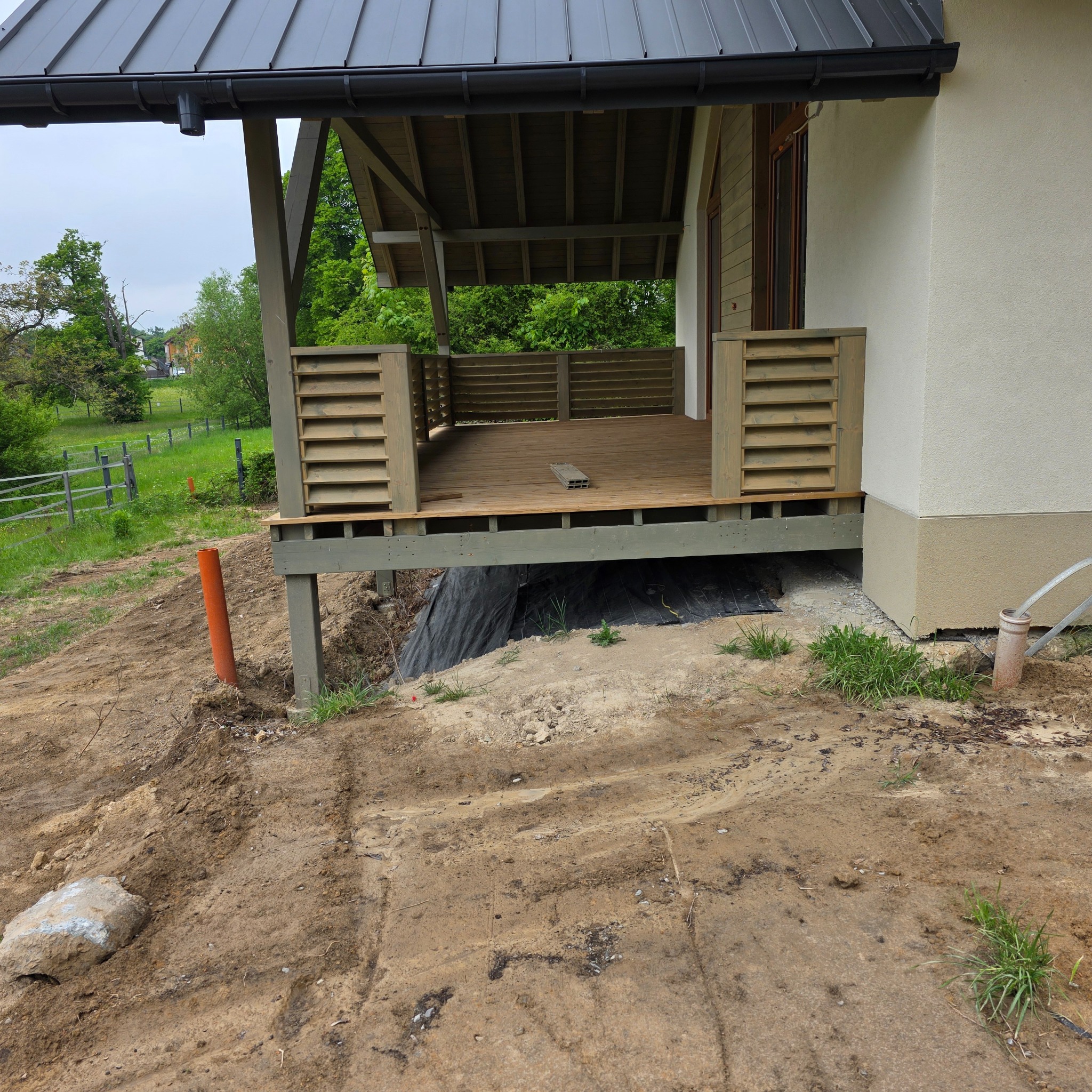 Drewniany taras z ażurowymi balustradami przy domu z jasną elewacją. Widoczne elementy konstrukcyjne i otoczenie w trakcie prac ziemnych.