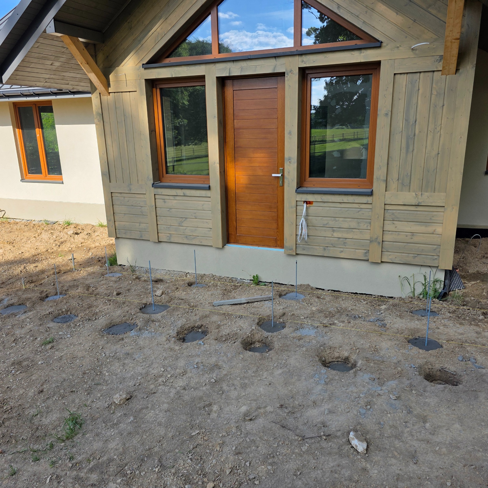 Fundamenty pod drewniany dom szkieletowy z widocznymi otworami i stalowymi prętami, teren przygotowany pod budowę. Widoczny fragment elewacji budynku.