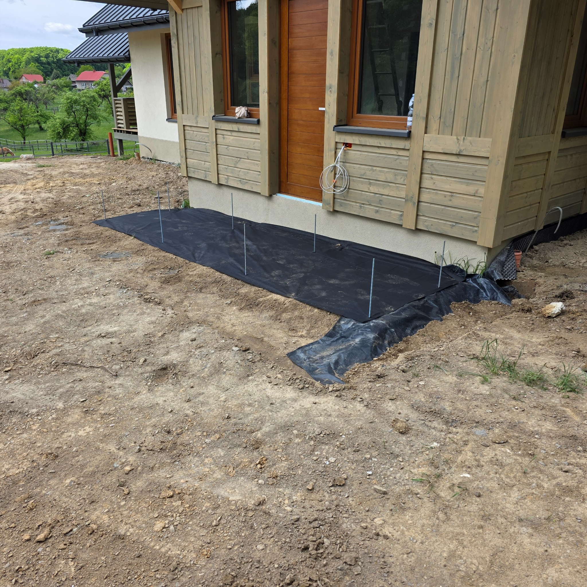Przygotowanie podłoża pod taras przy domu szkieletowym: czarna mata geowłókniny, metalowe pręty stabilizujące. Widoczny fragment elewacji z drewnianymi elementami.
