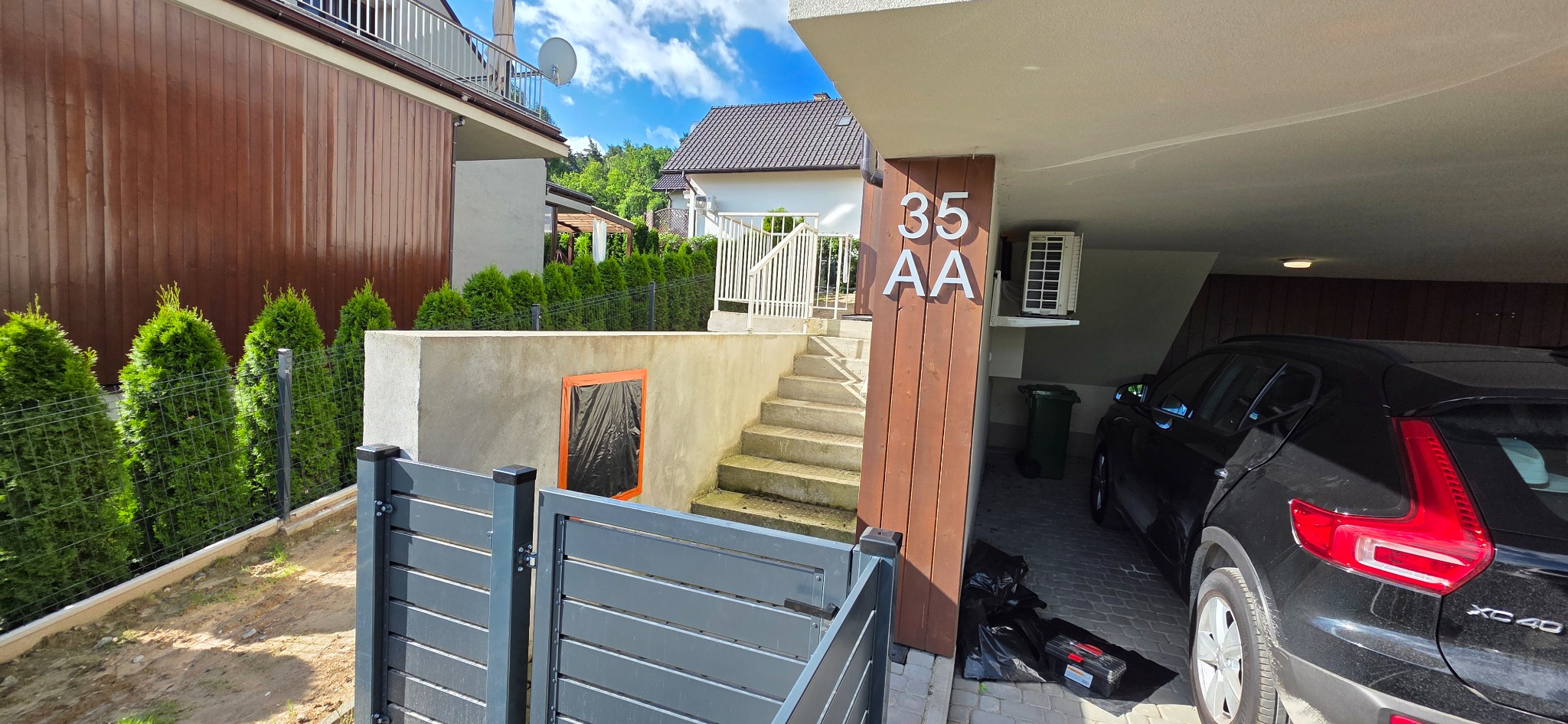 Dom z drewnianą elewacją i numerem 35 AA przy wjeździe do garażu. Widoczne schody, ogrodzenie, tuje i czarny samochód XC40.