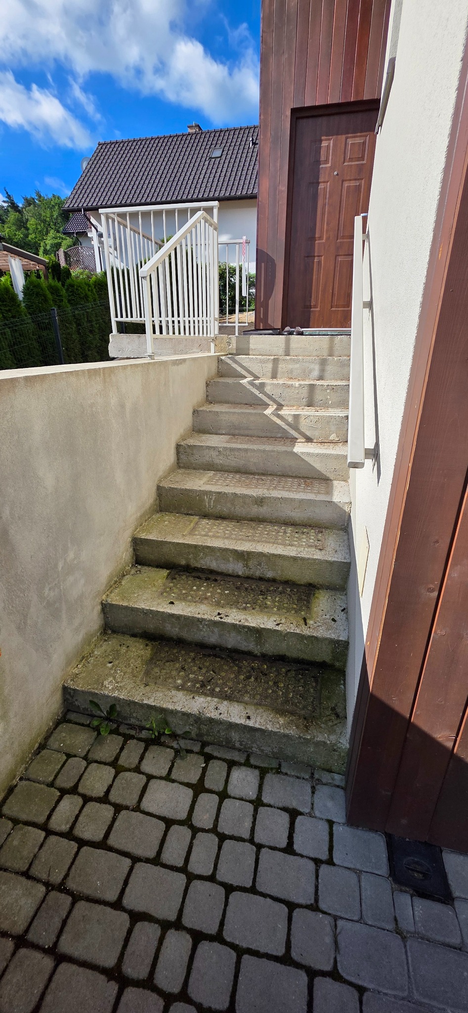 Betonowe schody zewnętrzne z białą balustradą prowadzą do brązowych drzwi wejściowych. Kostka brukowa u podstawy, błękitne niebo w tle.