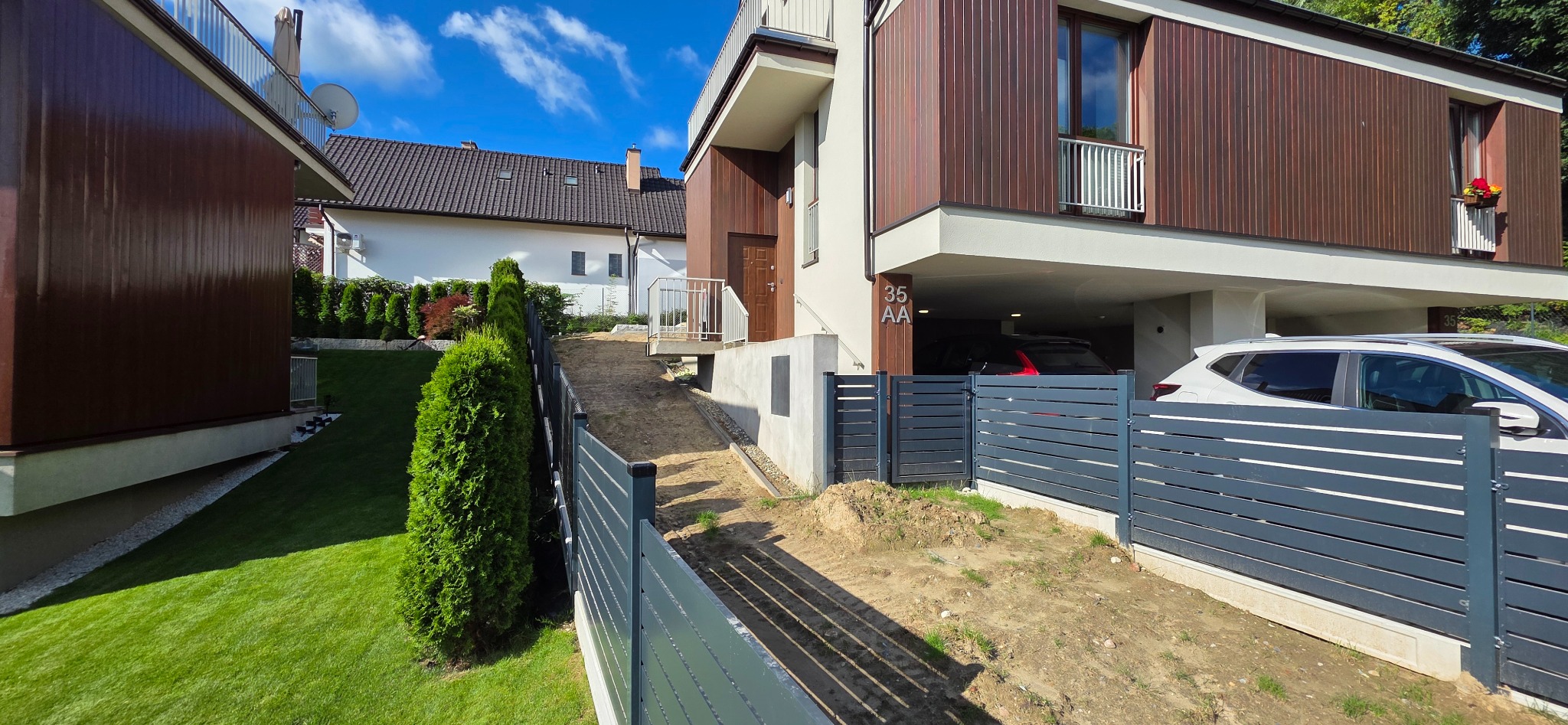 Nowoczesny dom z elewacją z paneli drewnopodobnych i szarą, poziomą zabudową ogrodzenia. Widoczny podjazd, zieleń i fragment sąsiedniego budynku.