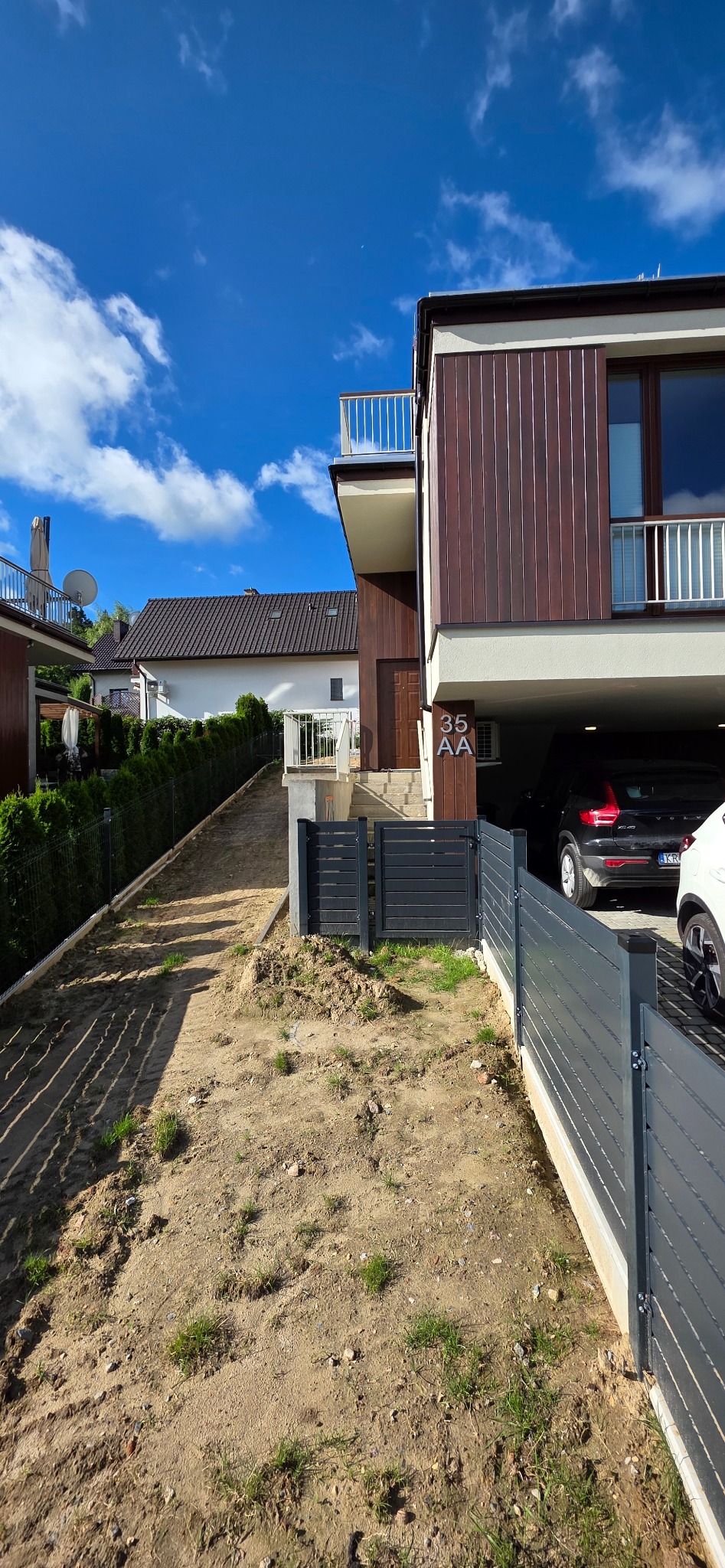 Nowoczesny dom z brązową elewacją i szarym ogrodzeniem panelowym, widok na podjazd i garaż z samochodami, błękitne niebo z chmurami w tle.