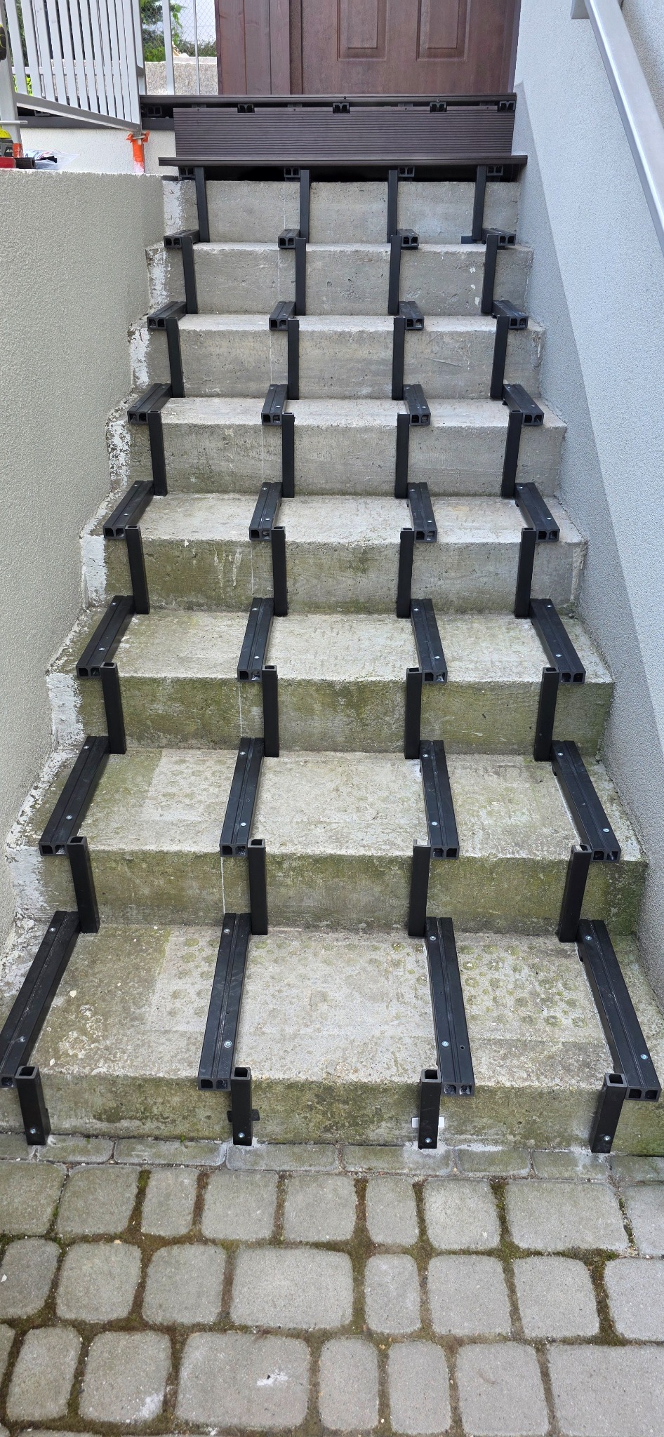 Betonowe schody z zamontowanymi elementami kompozytowymi, przygotowane do montażu desek. Widok z dołu na brązowe drzwi wejściowe.