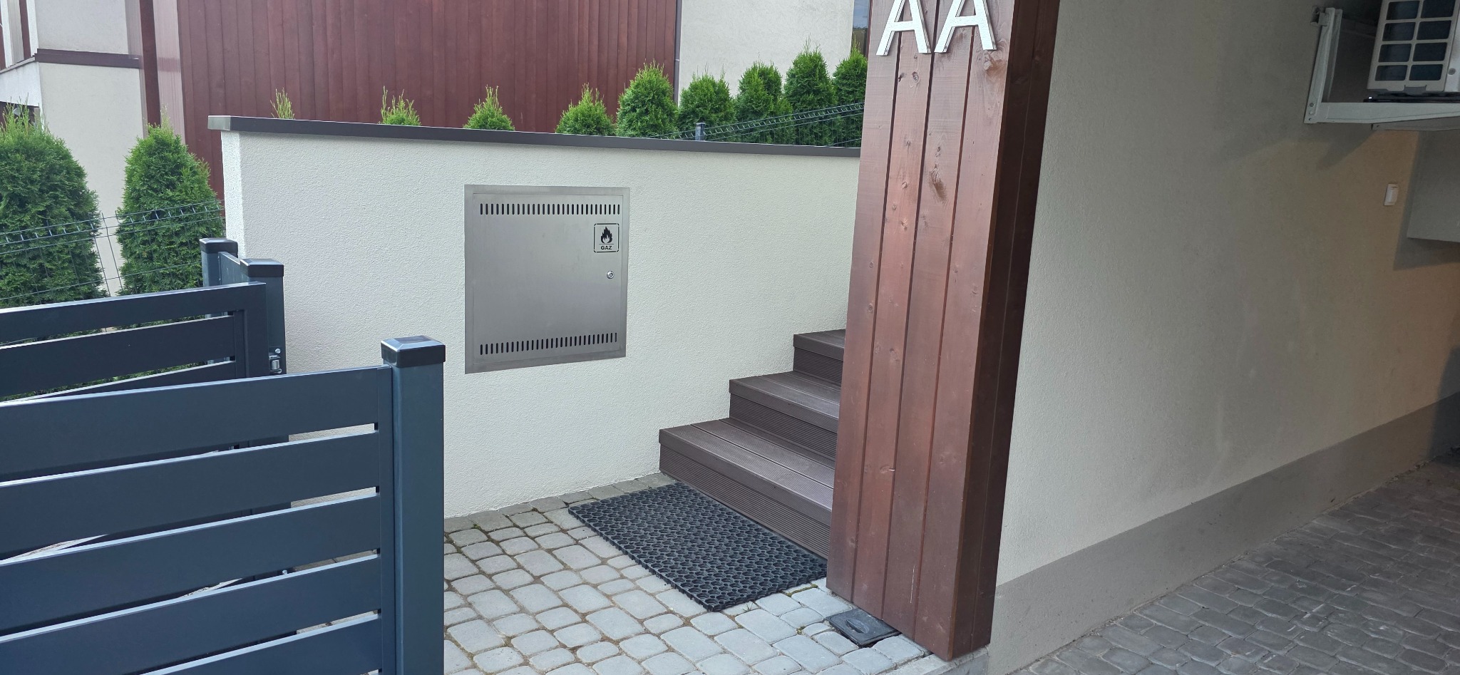 Schody z deski kompozytowej prowadzą do wejścia. Metalowa skrzynka gazowa na elewacji. Fragment szarego ogrodzenia. Na słupku drewnianym numer AA.