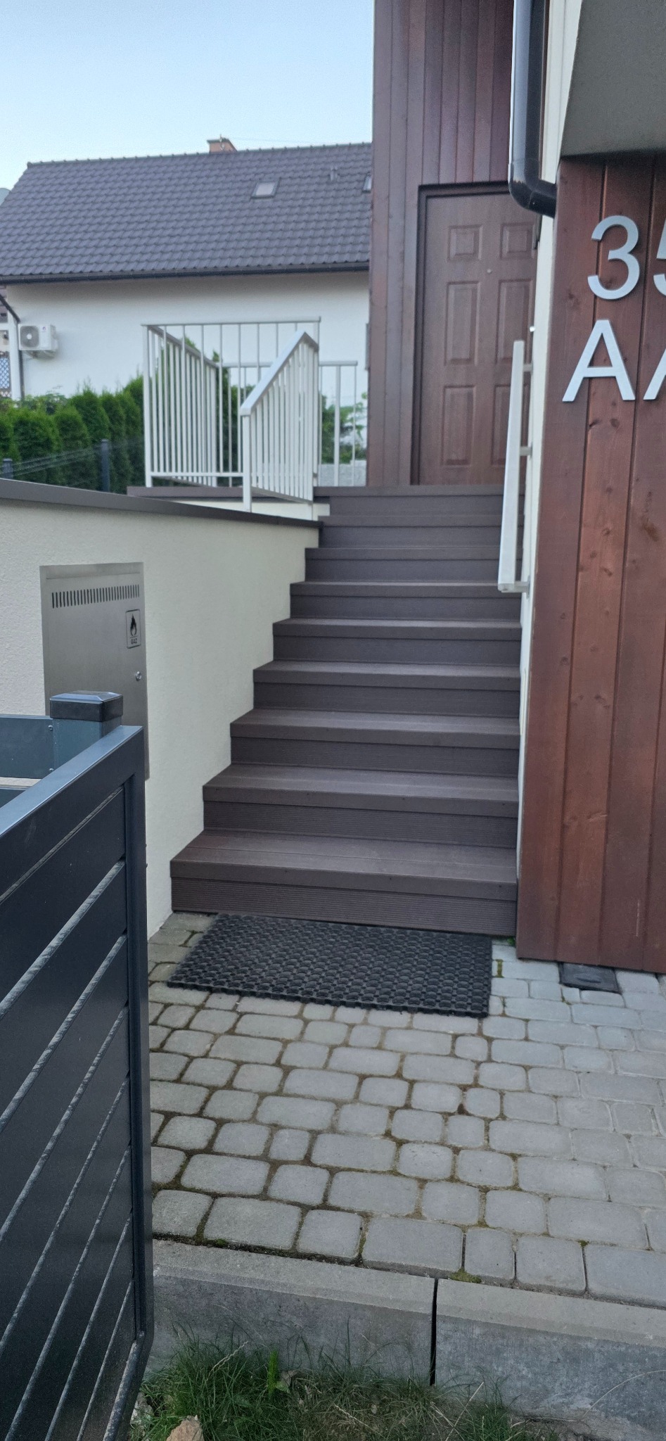 Schody wejściowe z brązowych desek prowadzące do drzwi domu. Widoczna wycieraczka i fragment chodnika z kostki brukowej. Po lewej stronie fragment metalowego ogrodzenia.