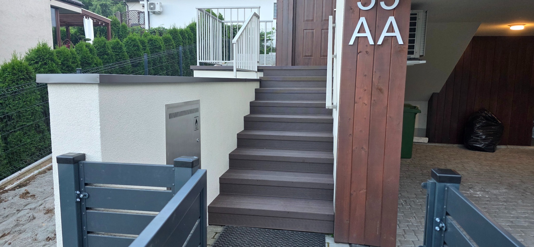 Schody z desek kompozytowych prowadzące do domu, z widocznym licznikiem gazu, szarą bramką i zielonymi tujami w tle. Oświetlenie naturalne.