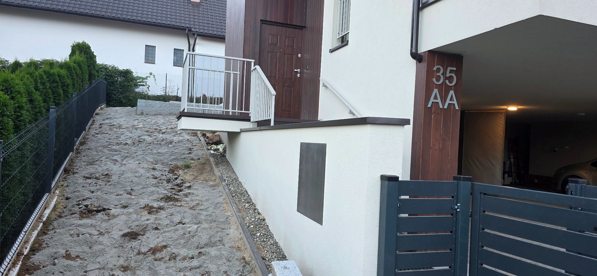 Nowoczesny dom z białymi ścianami i drewnianymi elementami wykończeniowymi, widoczny numer domu '35 AA', brama wjazdowa i fragment podjazdu.