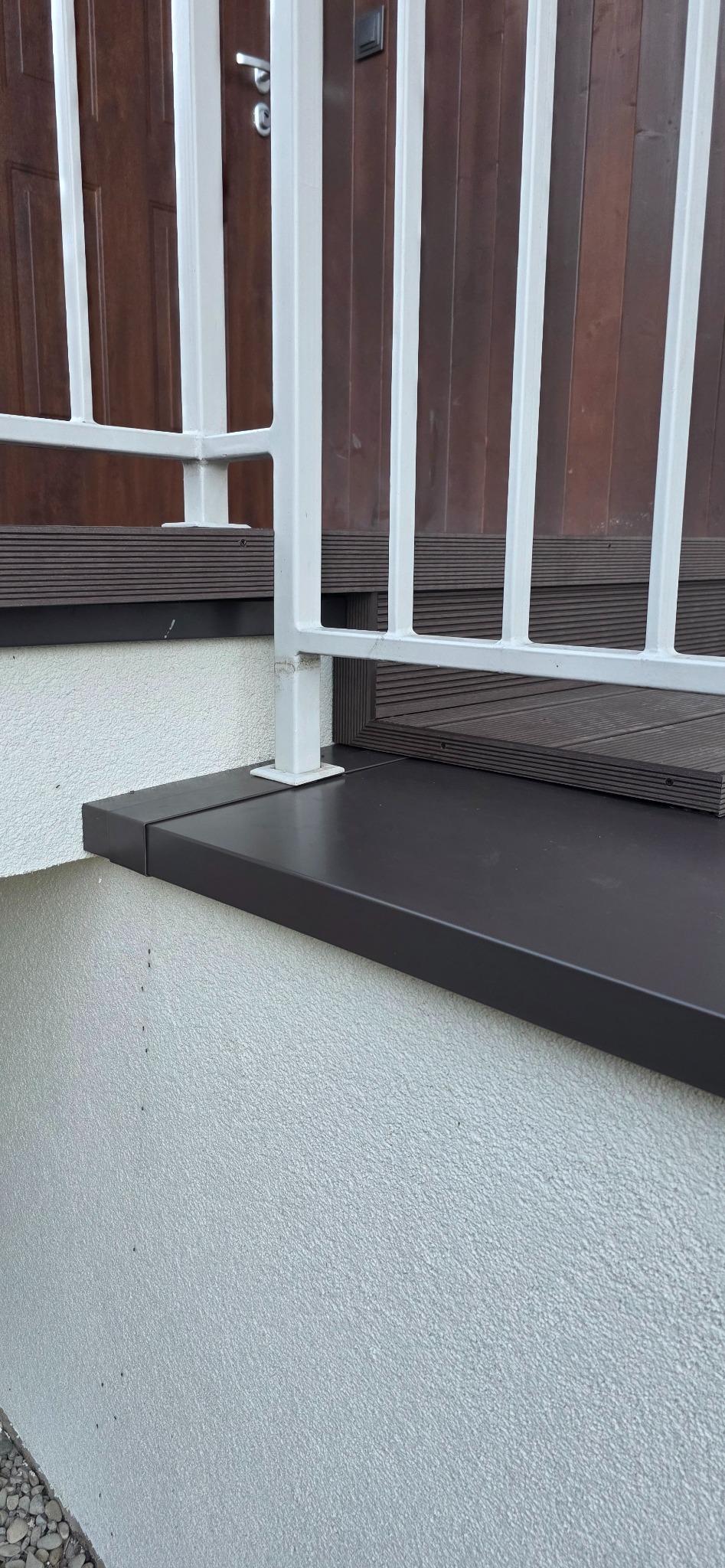 Detal wykończenia tarasu: deska kompozytowa w kolorze ciemnego brązu, biała balustrada i szara elewacja. Widoczny fragment drzwi wejściowych.