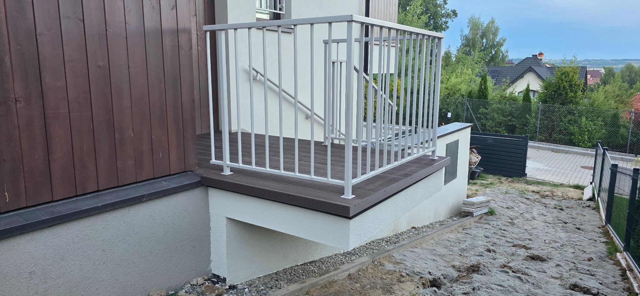 Nowoczesny taras z brązową deską kompozytową i białą balustradą, widok z boku. Na drugim planie zabudowa jednorodzinna i zieleń.