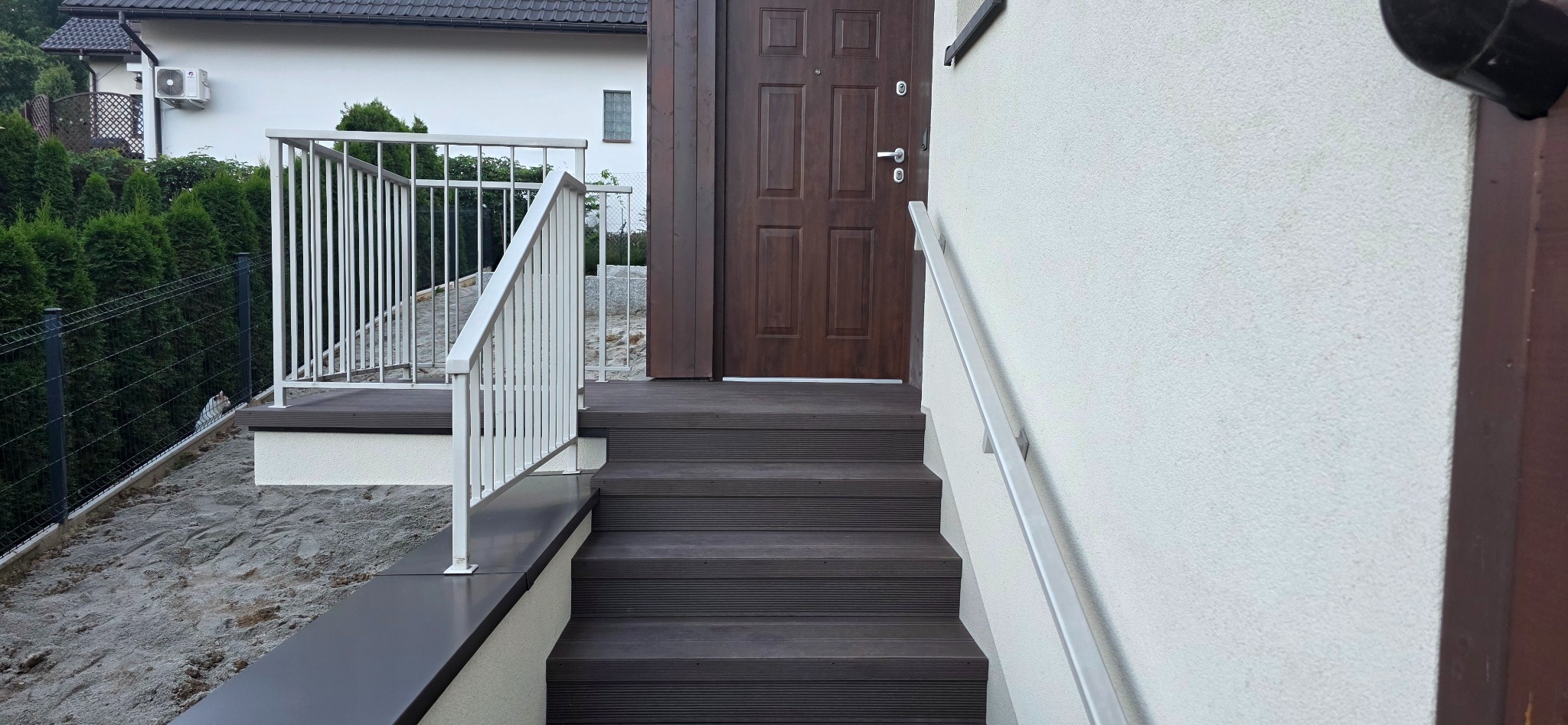Kompozytowe schody wejściowe z białą balustradą, prowadzące do brązowych drzwi w białym budynku. Widoczne elementy ogrodu i elewacji.