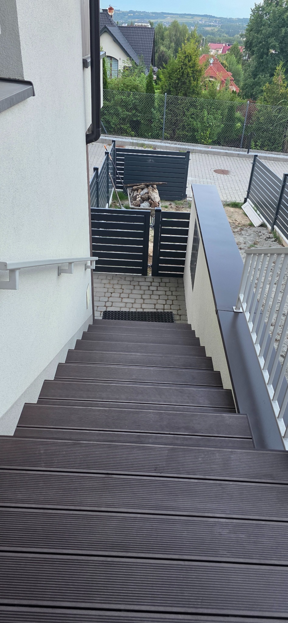 Schody z deski kompozytowej prowadzące do furtki z metalowymi panelami w nowoczesnym ogrodzie z widokiem na okolicę. Detal wykończenia.