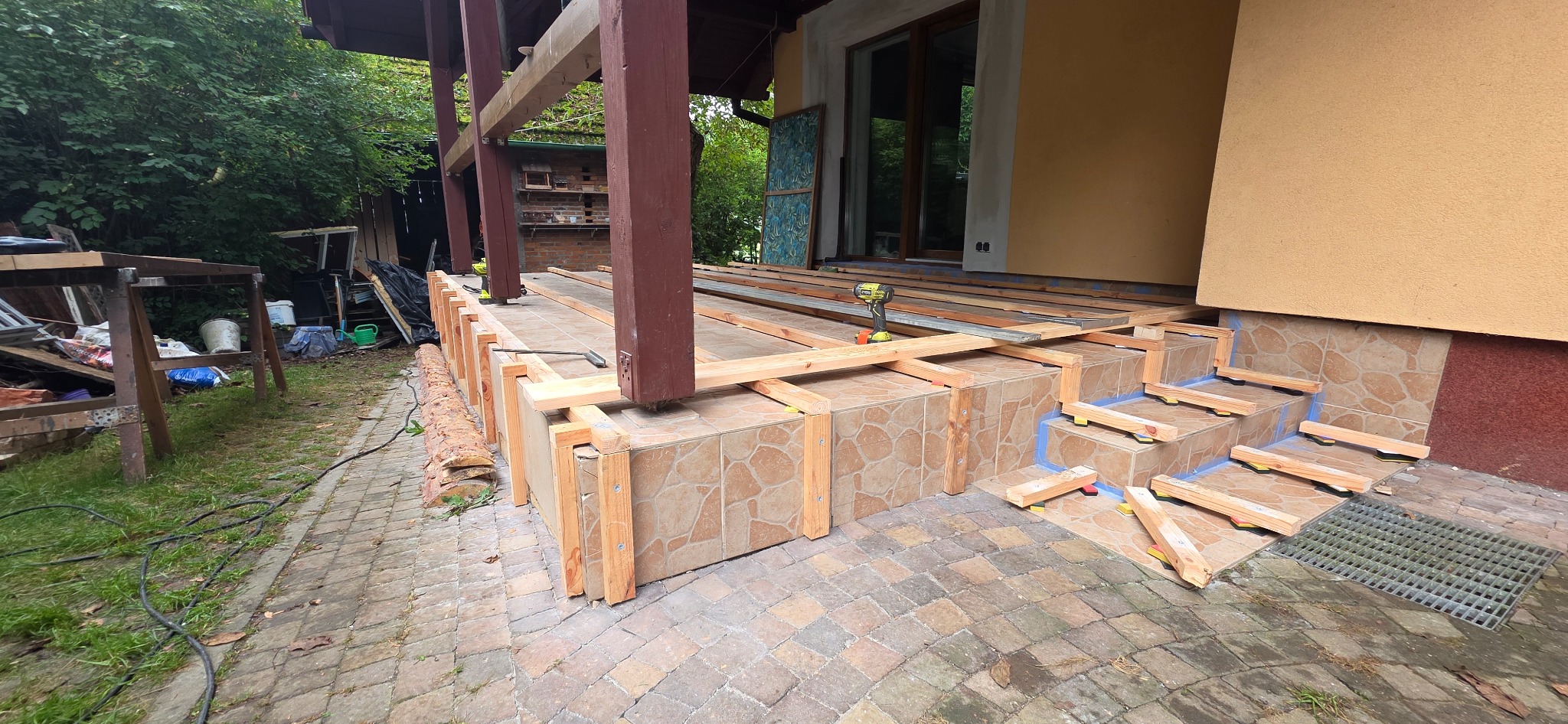 Budowa tarasu: drewniana konstrukcja na ceramicznych schodach, widoczny wkrętak akumulatorowy, fragment elewacji domu i otoczenie z zielenią.