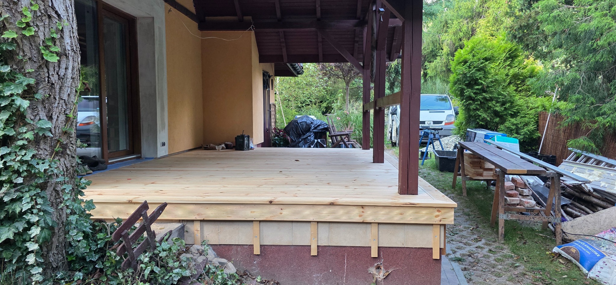 Jasny, drewniany taras przydomowy w trakcie budowy, z widoczną konstrukcją zadaszenia i elementami ogrodu w tle. Widoczne narzędzia i materiały budowlane.