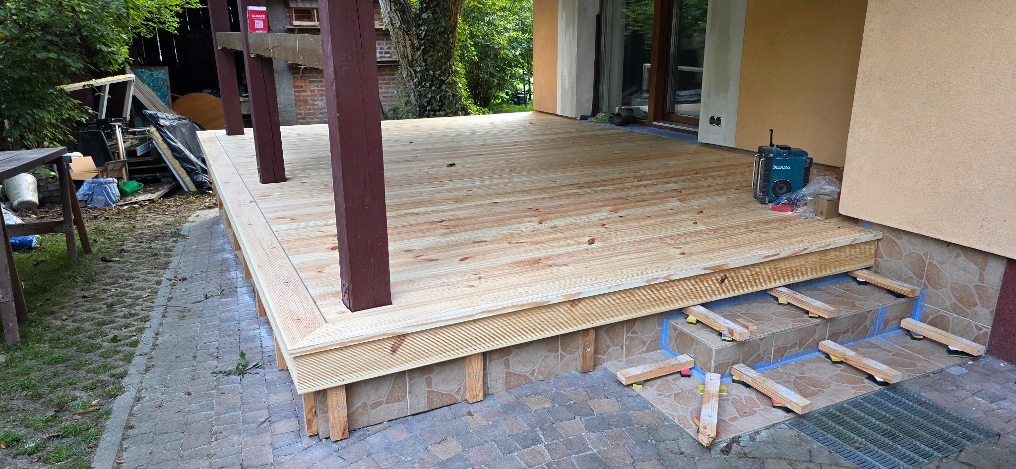 Świeżo wybudowany taras z jasnego drewna, z widocznymi podporami i narzędziem akumulatorowym Makita obok ściany budynku. Widok na schody z elementami poziomującymi.