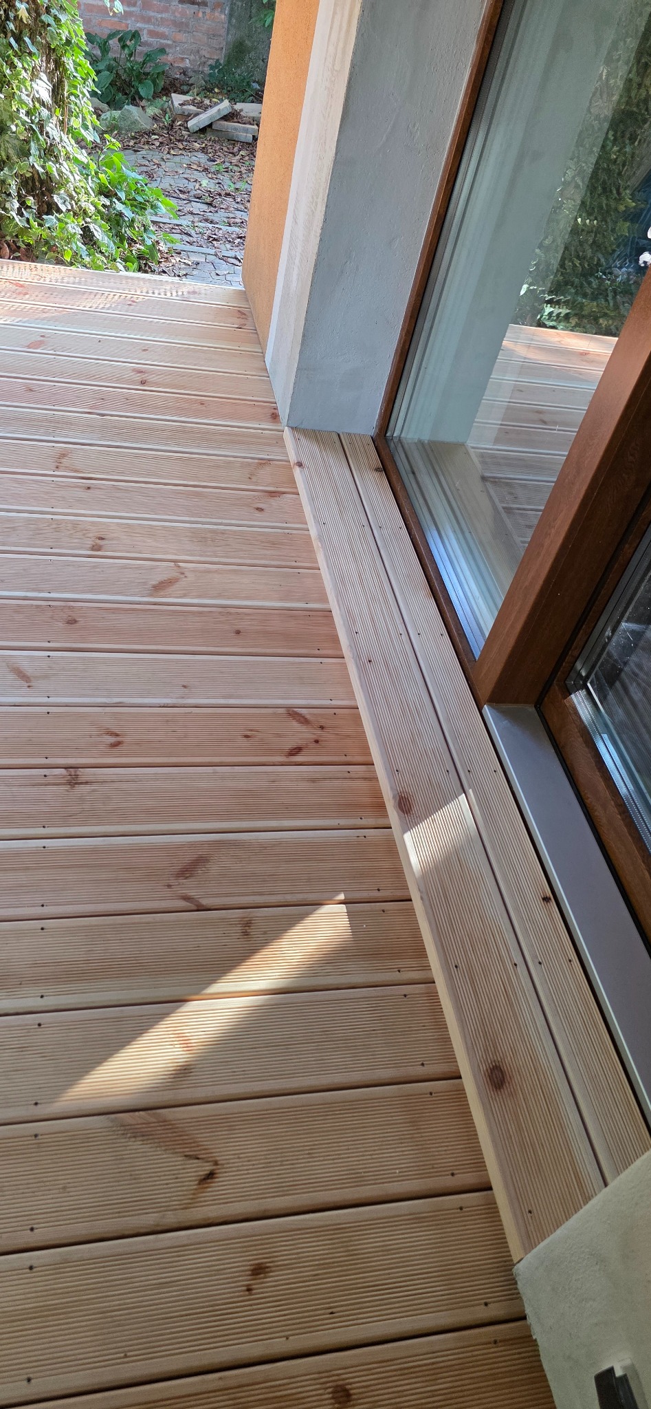 Jasny taras z desek, widok z góry, z subtelnym cieniem i widoczną strukturą drewna, sąsiadujący z oknem. Detal wykończenia i naturalne światło.