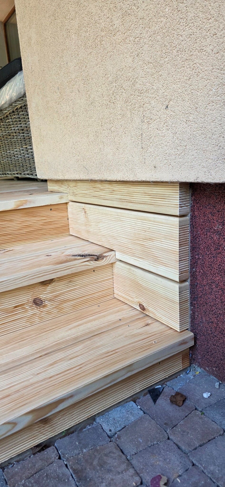 Detale drewnianych schodów zewnętrznych przylegających do ściany budynku z widoczną fakturą tynku. Widoczne kostki brukowe u podstawy schodów.