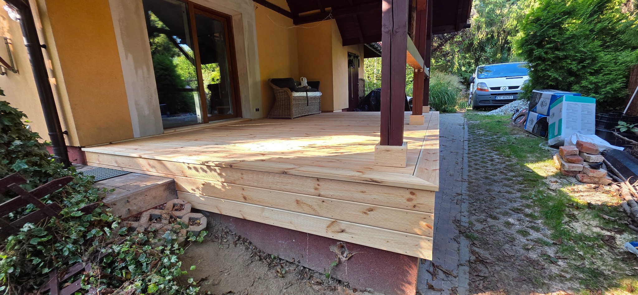 Jasny taras drewniany z widocznymi słojami drewna, częściowo zadaszony, z rattanowym siedziskiem w tle. Na zewnątrz zaparkowany biały samochód dostawczy.