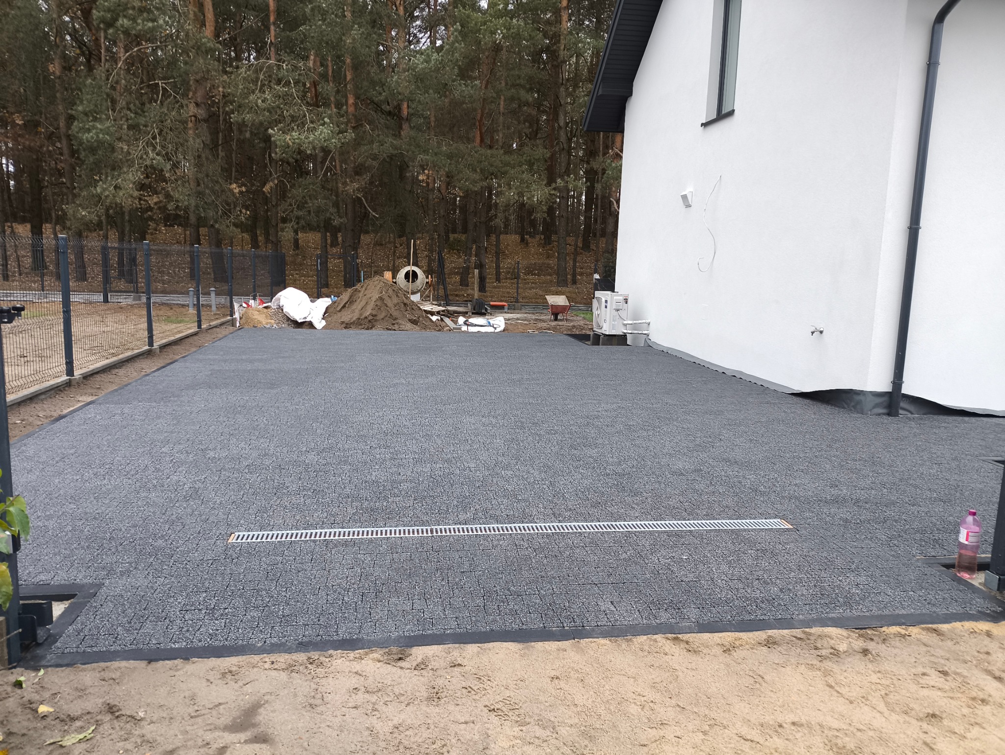 Ułożona szara nawierzchnia z kostki brukowej z odpływem liniowym, widoczna część domu z białą elewacją i fragment ogrodzenia panelowego w tle.