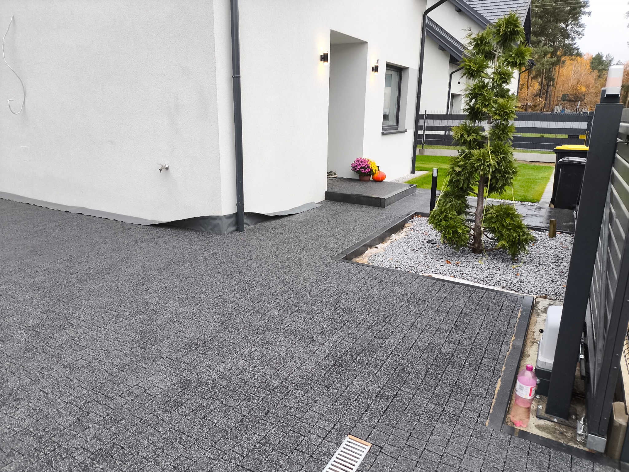 Nowoczesny podjazd z szarej kostki brukowej prowadzący do białego domu z ciemnymi akcentami, z widocznym systemem odprowadzania wody liniowego i elementami dekoracyjnymi przy wejściu.