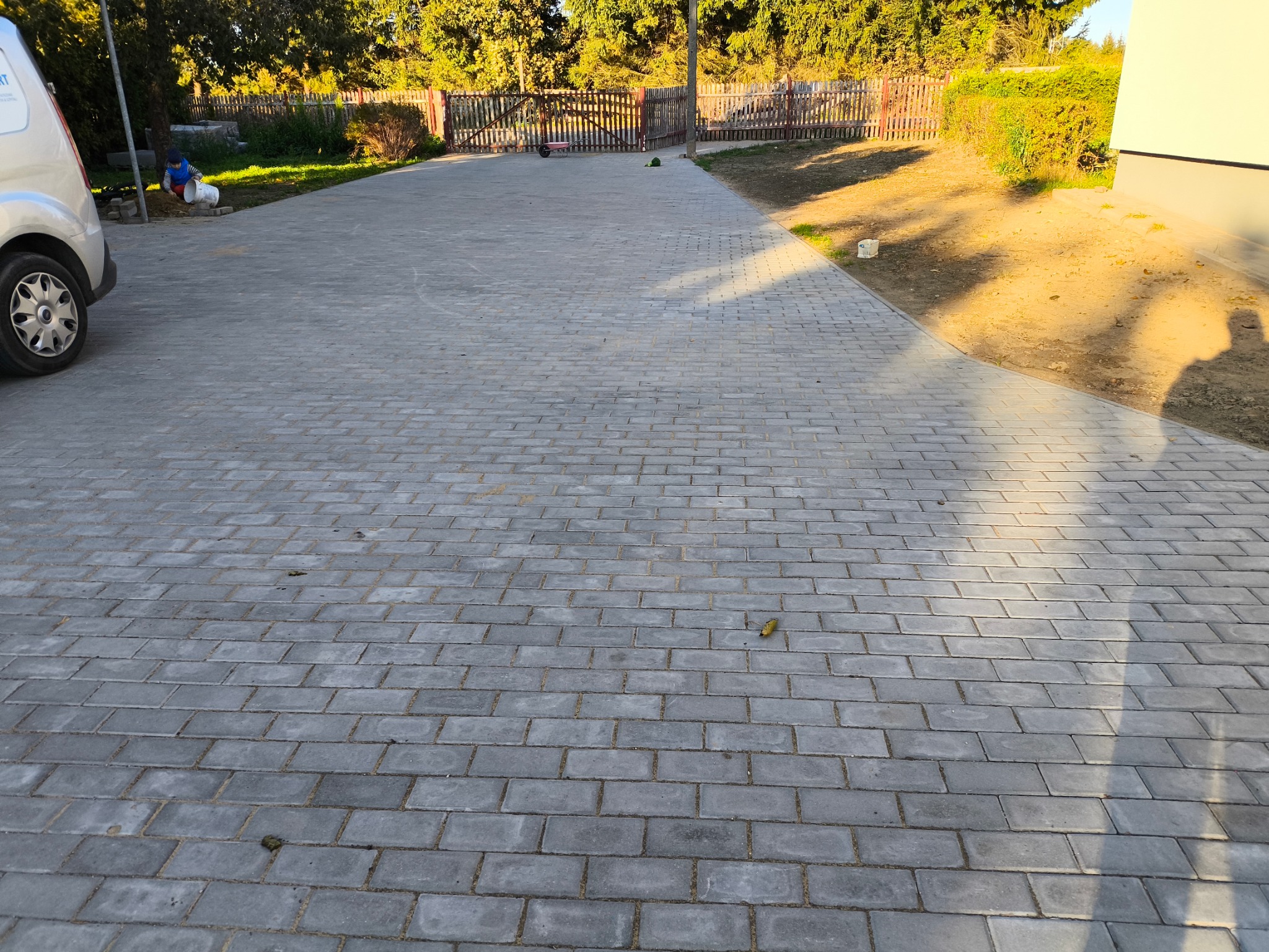 Szara kostka brukowa ułożona na podjeździe, w tle widoczny fragment ogrodzenia i zaparkowany samochód dostawczy.