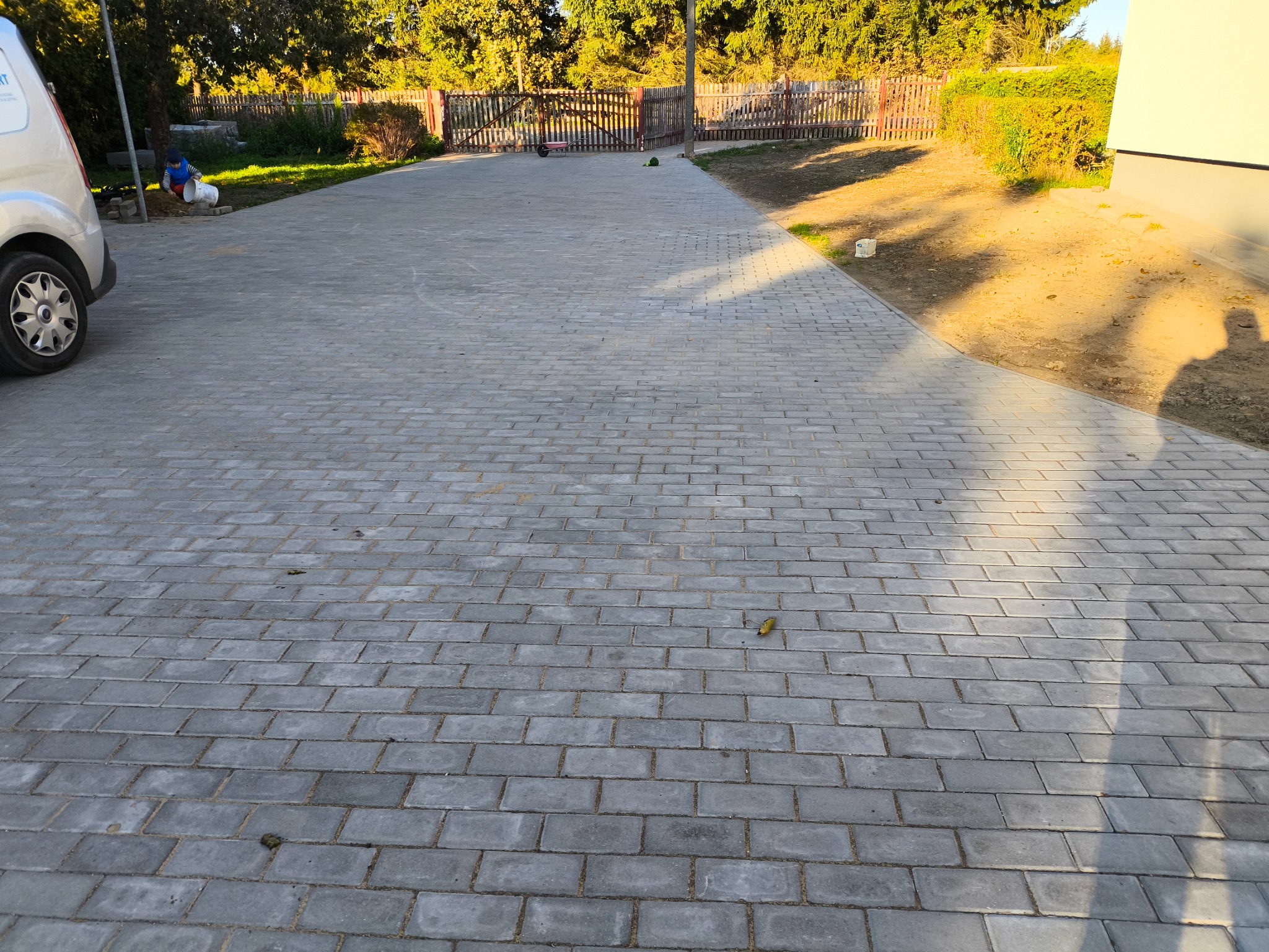 Szeroki, szary podjazd z kostki brukowej w kształcie prostokątów, zarośnięty delikatnie piaskiem, w tle ogrodzenie z drewnianych sztachet i częściowo zacieniony budynek. W lewym górnym rogu...