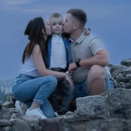 Rodzina: mama, tata i syn siedzą na kamiennym murku na tle miasta, rodzice całują syna w policzki, na murku widnieją napisy wykonane kredą.