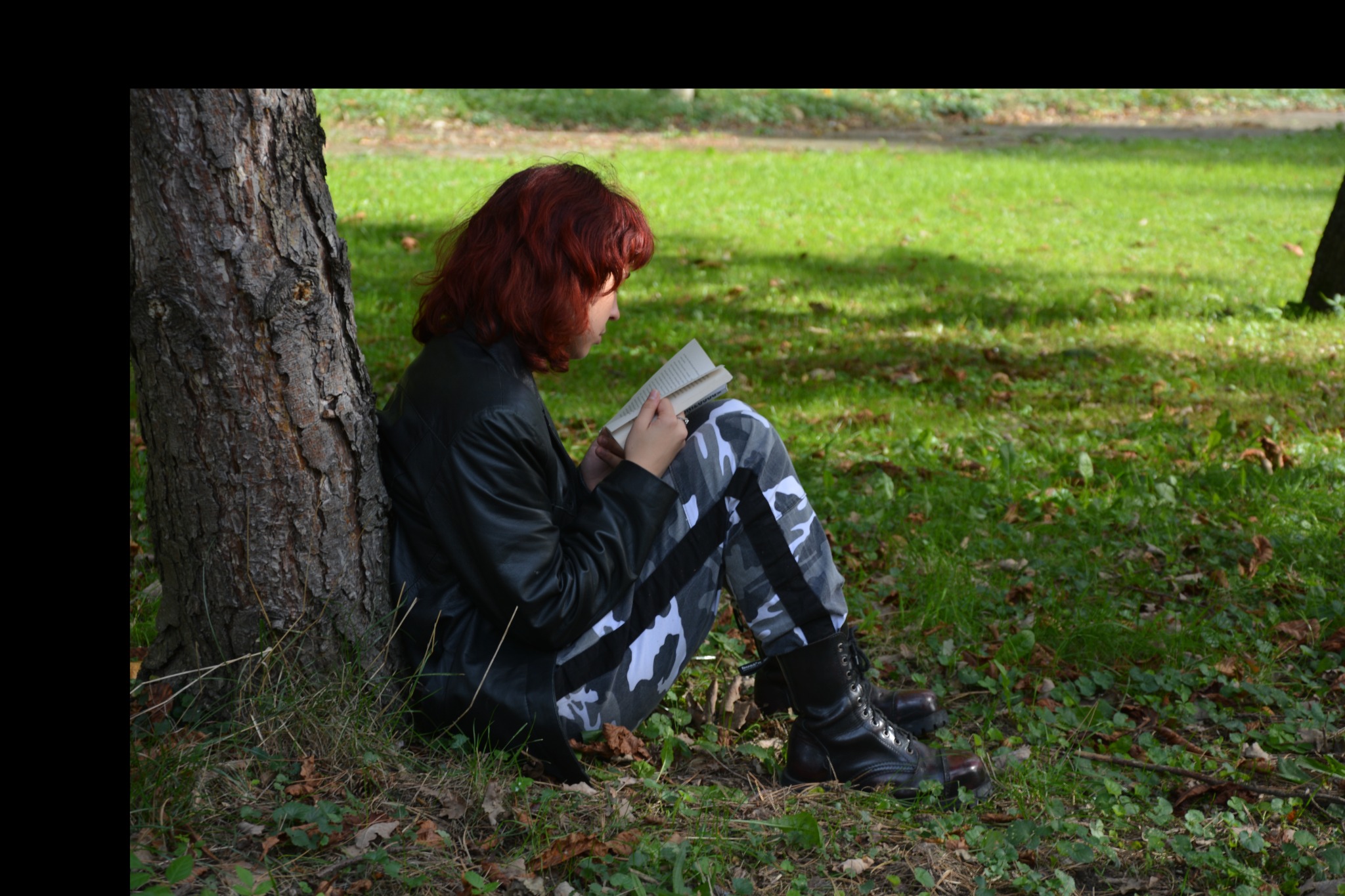 Kobieta z rudymi włosami w czarnej kurtce i wojskowych butach czyta książkę oparta o drzewo na trawie.