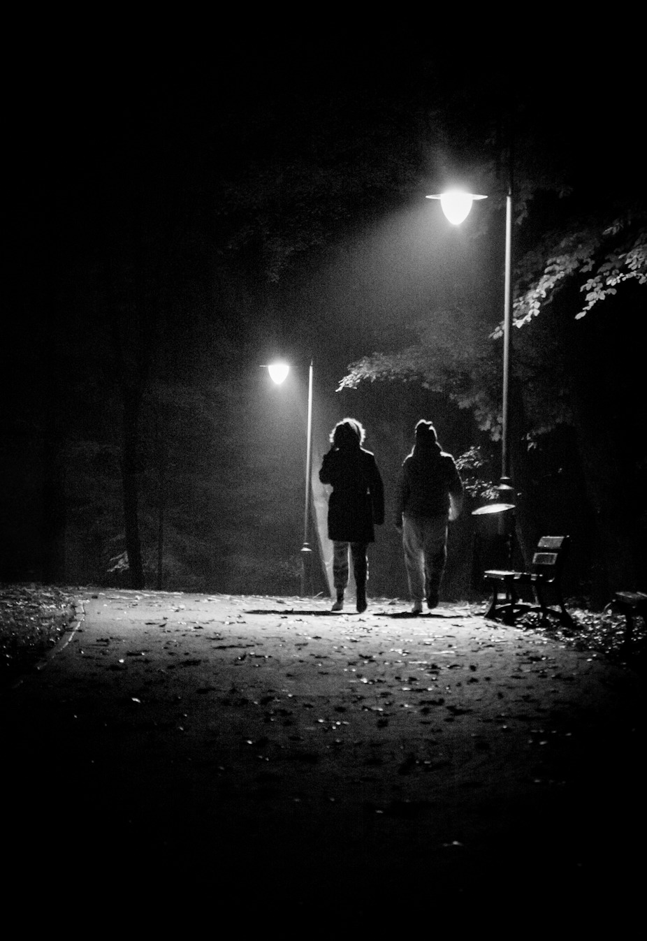 Dwie osoby oddalają się alejką w parku nocą, oświetloną przez latarnie, widoczne ławki i drzewa w tle, czarno-biała fotografia