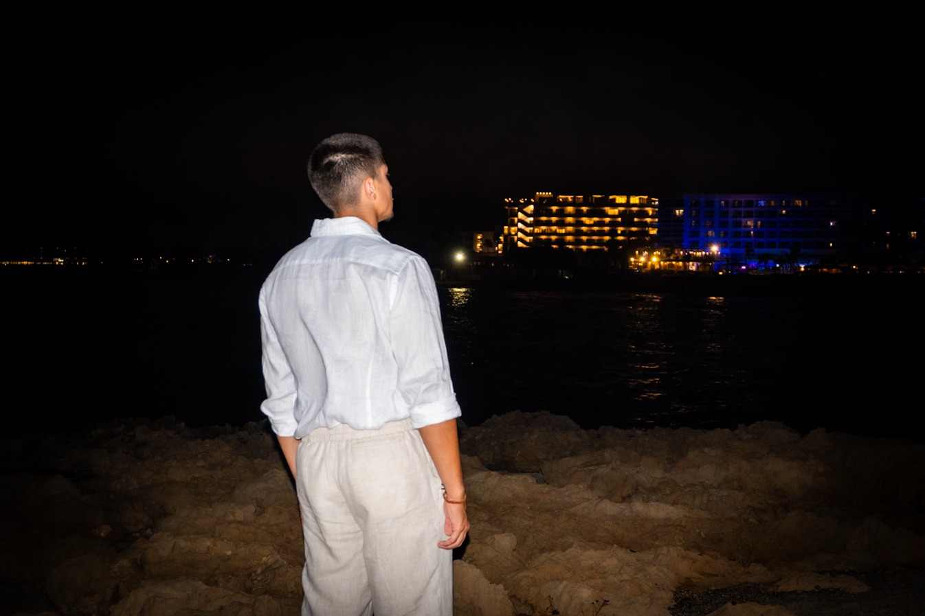 Mężczyzna w białej koszuli i lnianych spodniach stoi tyłem na skalistym brzegu, w oddali widoczne oświetlone budynki hotelowe nocą.