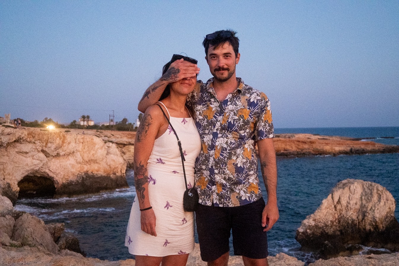 Para narzeczonych na tle skalistego wybrzeża z tatuażami, kobieta zasłania oczy okularami przeciwsłonecznymi.