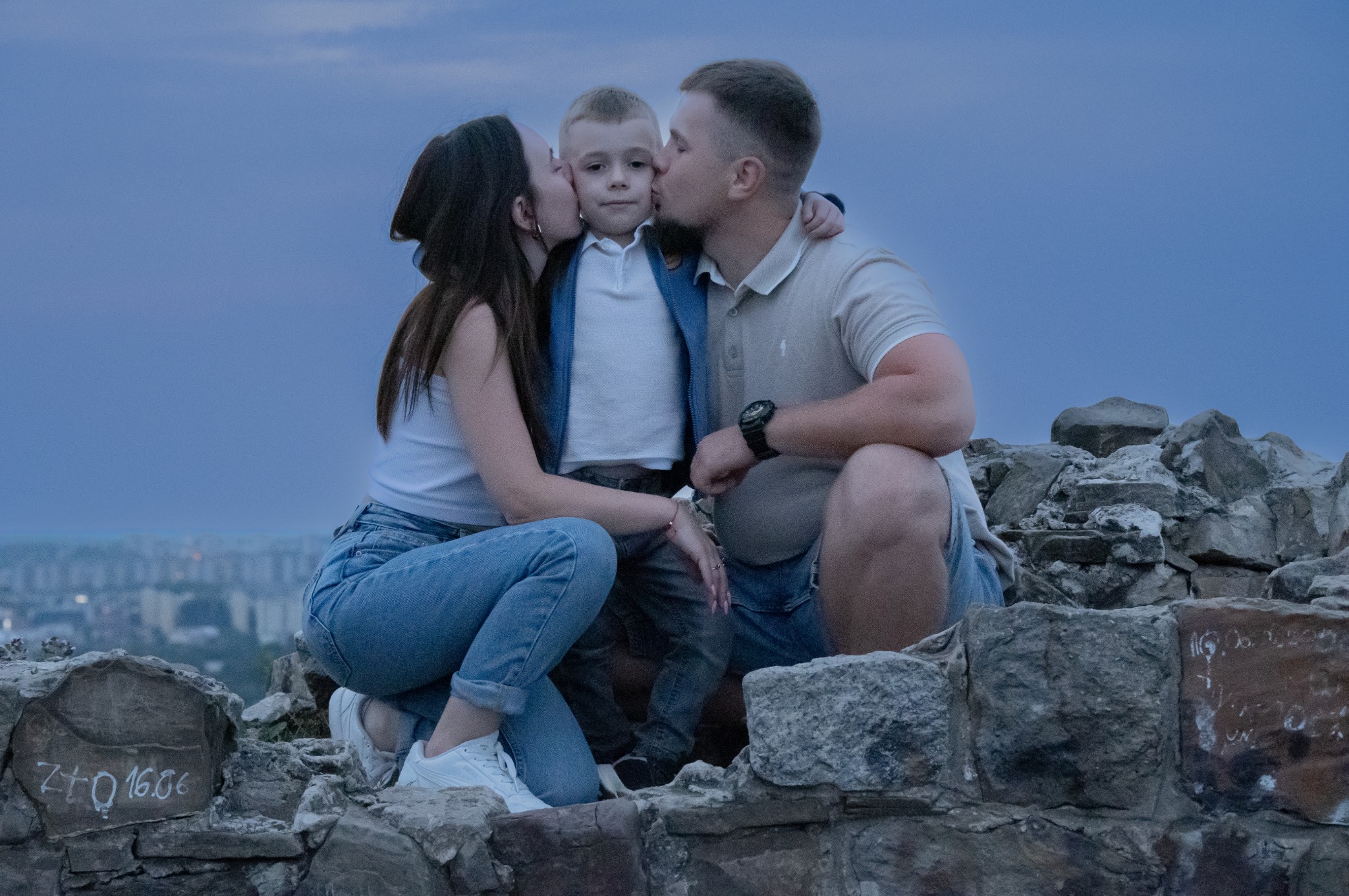 Rodzina: mama, tata i syn siedzą na kamiennym murku na tle miasta, rodzice całują syna w policzki, na murku widnieją napisy wykonane kredą.