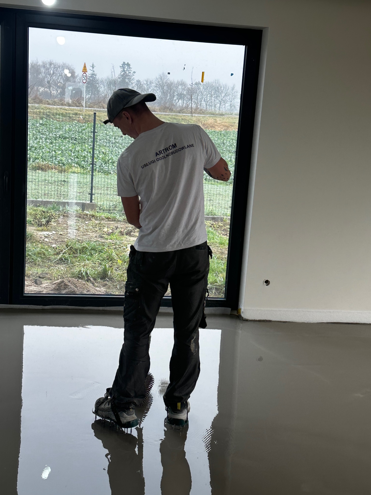 Pracownik w butach z kolcami rozprowadza wylewkę samopoziomującą w jasnym pomieszczeniu z dużym oknem. Widok z tyłu, logo firmy na koszulce.