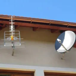 Dwie różne anteny zamontowane na elewacji domu: telewizyjna kierunkowa i satelitarna, pod brązowym dachem z drewnianą podbitką.