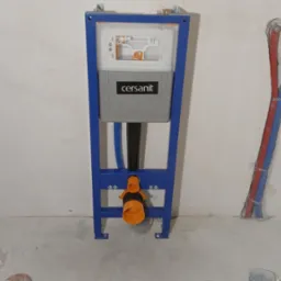 Stelaż podtynkowy Cersanit z widocznymi przyłączami wodnymi (czerwony i niebieski) w surowym stanie łazienki przed wykończeniem.