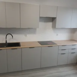 Nowoczesna, szara zabudowa kuchenna z drewnianym blatem i czarnym zlewem. Na blacie płyta indukcyjna. Białe płytki na ścianie z delikatnym wzorem.