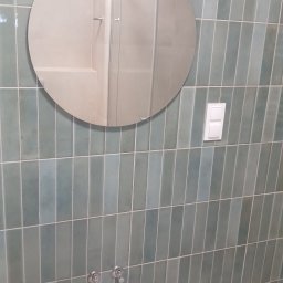 Kamil Pawłowski - Łazienka po remoncie: pionowe, turkusowe płytki, okrągłe lustro odbijające kabinę prysznicową, biały włącznik światła i bateria umywalkowa. Minimalistyczny design.