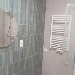 Kamil Pawłowski - Wykończona łazienka z pionowymi, turkusowymi płytkami, okrągłym lustrem, białym grzejnikiem drabinkowym i szklaną kabiną prysznicową. Minimalistyczny design.