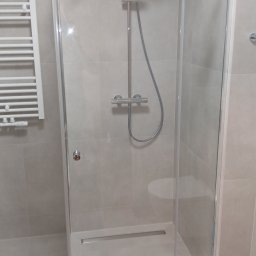 Kamil Pawłowski - Nowoczesna kabina prysznicowa z deszczownicą i odpływem liniowym, wykończona w jasnych, neutralnych barwach. Widoczny fragment grzejnika drabinkowego.