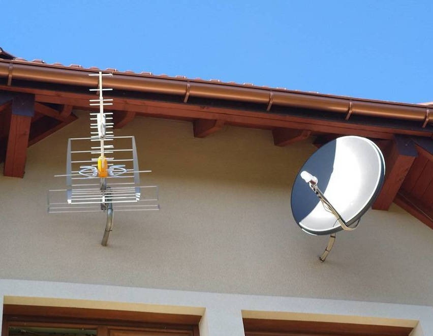 Dwie różne anteny zamontowane na elewacji domu: telewizyjna kierunkowa i satelitarna, pod brązowym dachem z drewnianą podbitką.