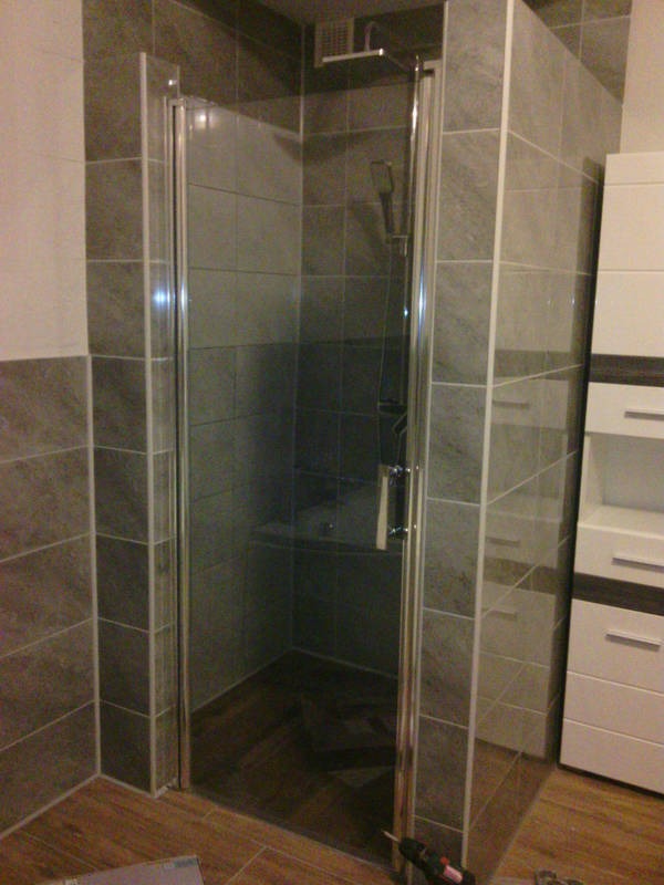 Szklana kabina prysznicowa z chromowanymi profilami, zamontowana w łazience wyłożonej szarą płytką imitującą kamień, widoczna częściowo biała szafka z szufladami, na podłodze brązowe płytki, w rogu...