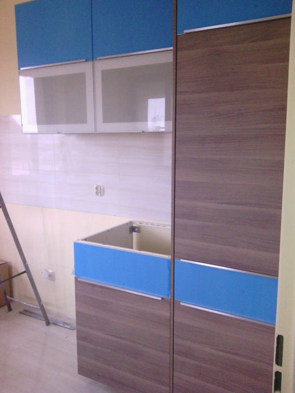 Wnętrze kuchni w trakcie urządzania: widoczne szafki górne z niebieskimi frontami i przeszklonymi drzwiczkami, dolne szafki z frontami w odcieniach brązu, brak blatu, drabina oparta o ścianę...