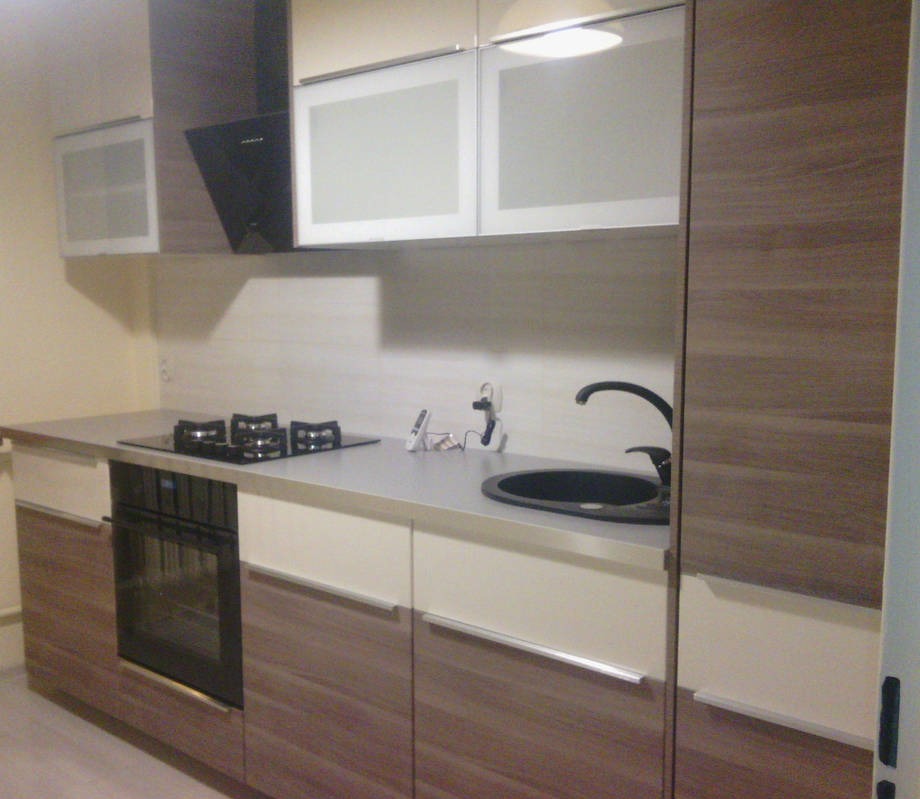 Nowoczesna kuchnia z dwukolorowymi frontami szafek (białe i drewnopodobne), czarnym zlewem i płytą grzewczą oraz okapem w ciemnym kolorze. Widoczne są również górne szafki z matowymi, jasnymi...