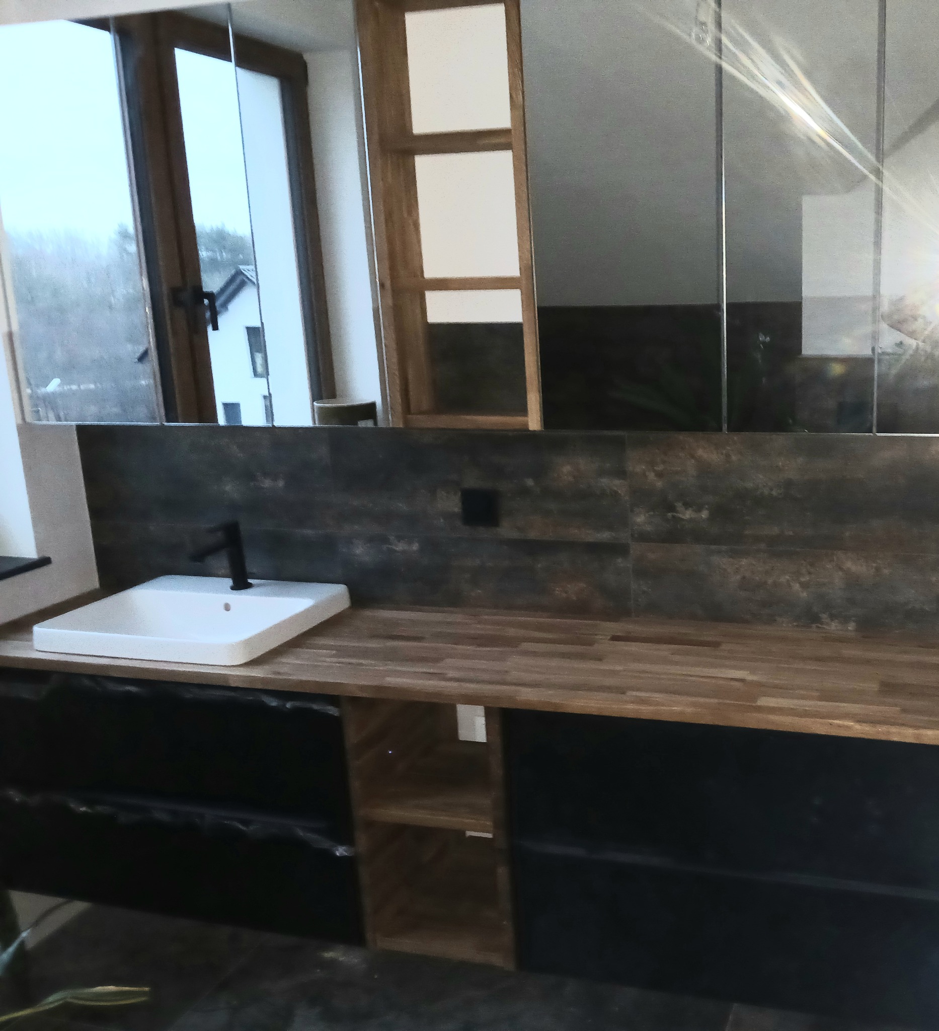 Nowoczesna łazienka z drewnianym blatem, białą umywalką z czarną baterią i lustrzanymi szafkami, widok na okno z domem w tle.