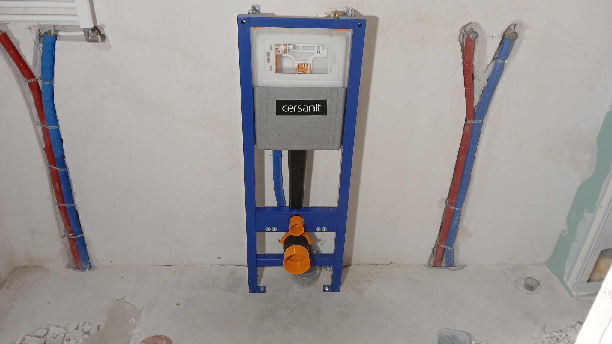 Stelaż podtynkowy Cersanit z widocznymi przyłączami wodnymi (czerwony i niebieski) w surowym stanie łazienki przed wykończeniem.