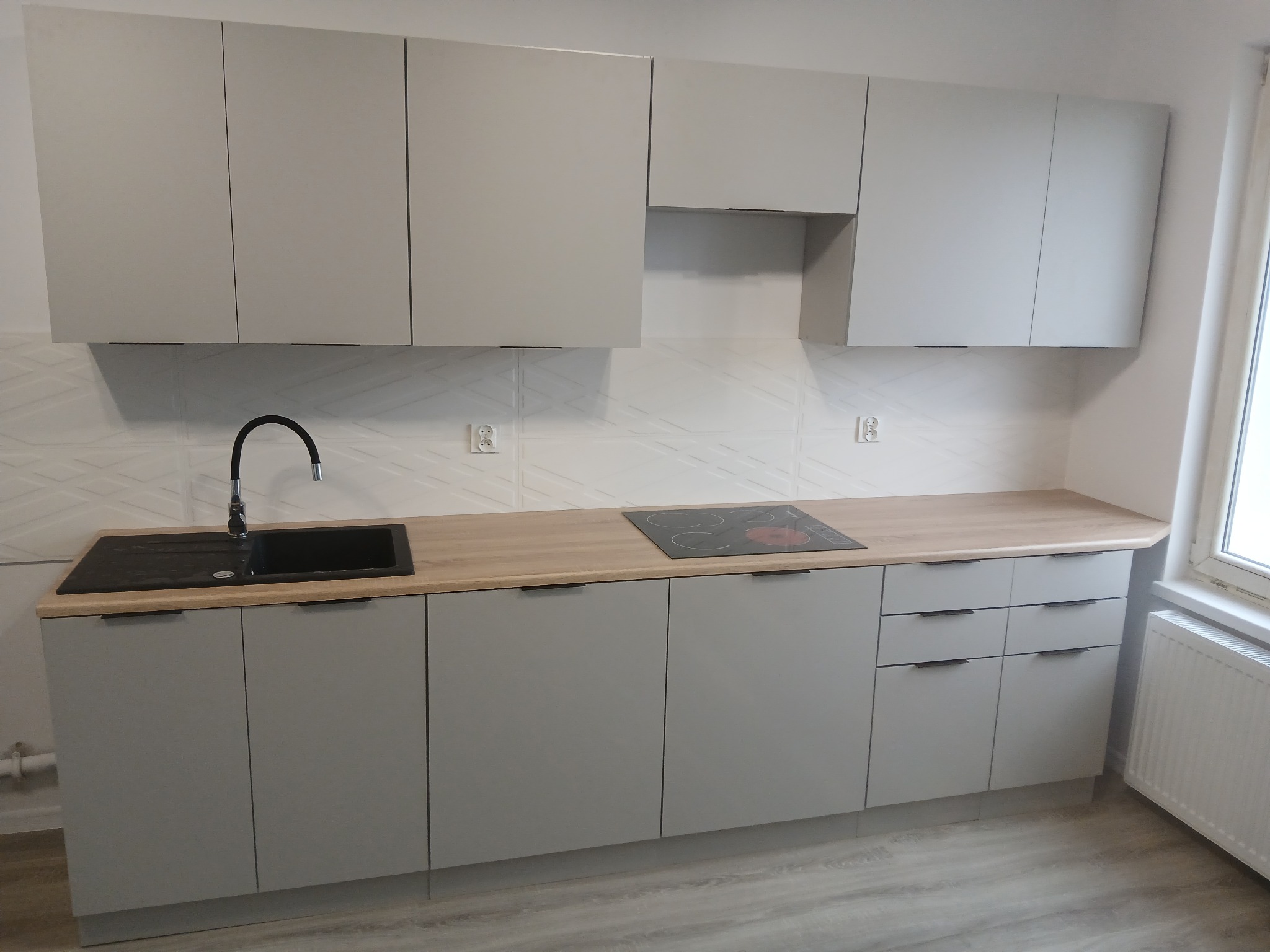 Nowoczesna, szara zabudowa kuchenna z drewnianym blatem i czarnym zlewem. Na blacie płyta indukcyjna. Białe płytki na ścianie z delikatnym wzorem.