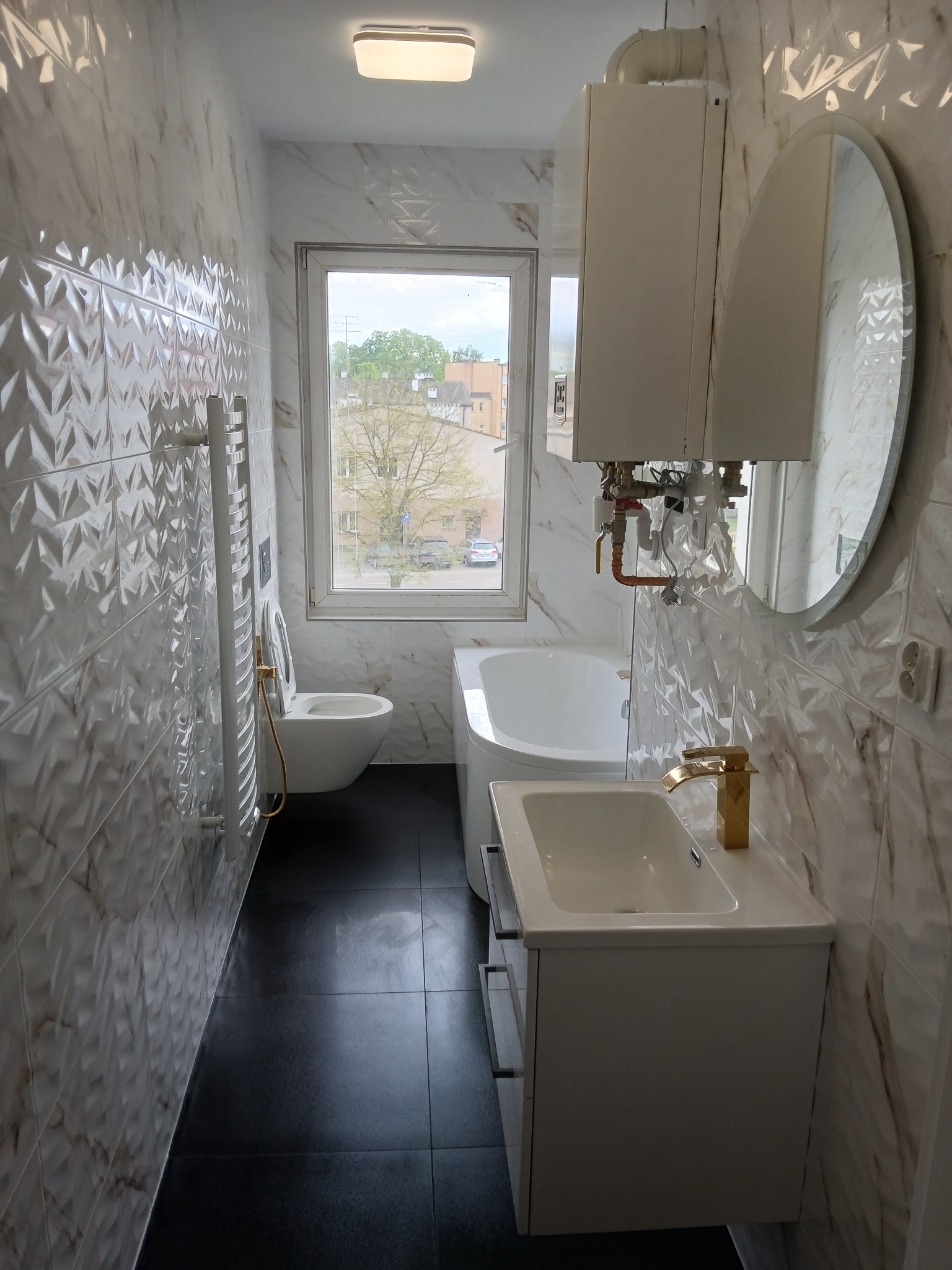 Wykończona łazienka z biało-marmurkowymi płytkami 3D, czarną podłogą i złotą armaturą. Widoczne WC, umywalka z szafką, grzejnik i bojler.