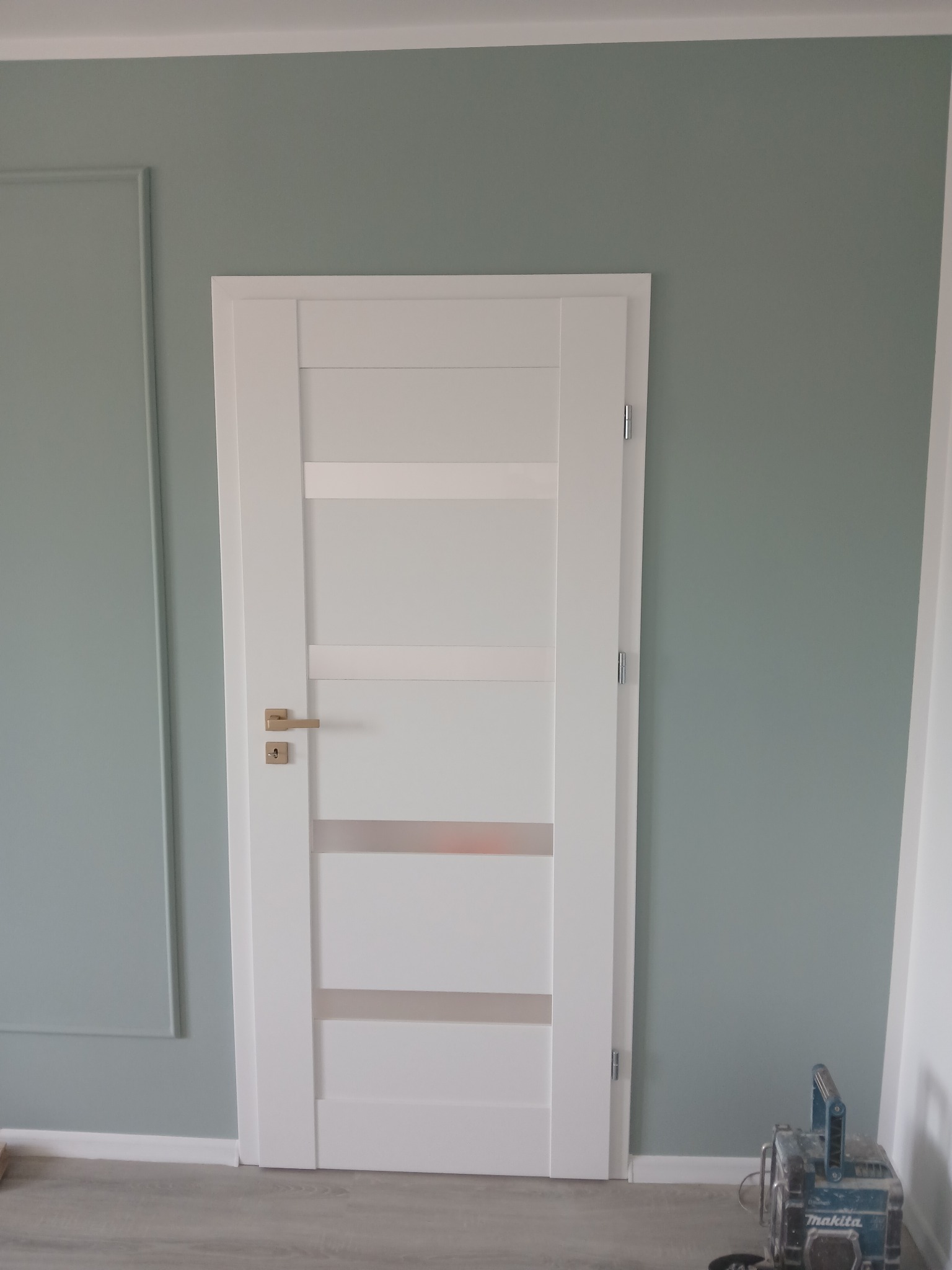 Białe, nowoczesne drzwi wewnętrzne z przeszkleniami, zamontowane w pokoju z szarozielonymi ścianami i listwą dekoracyjną. Na podłodze widoczny sprzęt budowlany.