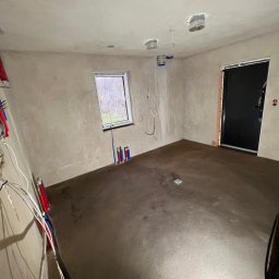 JS INVEST JAKUB DUNST - Wnętrze w trakcie prac: wylana posadzka, surowe ściany, instalacje elektryczne i hydrauliczne. Widoczne okno z widokiem na zewnątrz, czarne drzwi.