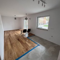 JS INVEST JAKUB DUNST - Jasne, przestronne wnętrze po częściowym remoncie: parkiet dębowy ułożony obok szarych płytek. Niebieska taśma oddziela powierzchnie. Widoczne lampy i okno z widokiem na miasto.